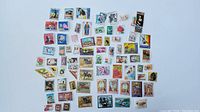 Top view of a diverse assortment of approximately 70 postage stamps, showing various designs, colors, and sizes spread out on a white surface.