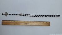 Full length view of black bead rosary necklace with sterling silver cross and pendant alongside ruler for size reference.