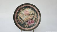 Front view of the circular ceramic plate displaying hand painted peacock and floral design with green background and pink/gold accents.