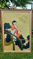 Full view of the framed painting showing the woman seated on tree branch playing flute, background with tree and foliage they are sitting on.