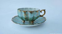 Blue lustreware teacup with scalloped shape, gold handles and three small gold feet, iridescent finish, sitting on matching saucer with scalloped edges and gold decoration.