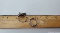Two silver rings shown next to a ruler for size reference, one with double mounted stones and one plain band with pattern.