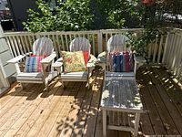 Three white Adirondack style patio chairs with mismatched cushions and a matching small side table on wooden deck