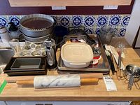 Wide view of bakeware and kitchen tools on countertop, includes springform pans, loaf pans, muffin tin, rolling pin, mixing bowl, decorating bag, and utensils.