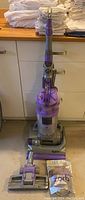 Dyson DC14 upright vacuum cleaner standing on the floor against cabinetry, main body purple and gray with clear dust bin.