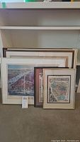 Photo showing six framed historical maps stacked against a wall, featuring different frame colors and styles.