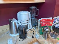 Photo shows white Kitchenaid drip coffee maker, stainless steel electric kettle, black coffee grinder, blender, and coffee accessories on counter.