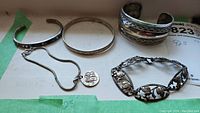 Five-piece sterling silver jewelry lot displayed on white paper with green tape, including chain necklace with pendant, three bracelets, and one cuff.