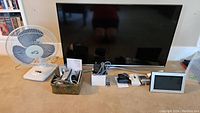 Samsung 40-inch flat screen TV, Panasonic cordless phones with docking stations in a basket, table fan, two digital clocks, Apple TV box visible.