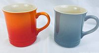 Two Le Creuset mugs side by side; one orange gradient, one blue, showing front and handles