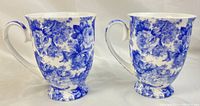 Two blue and white floral mugs side by side, front view, showing detailed floral pattern and curved handles.