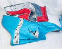 Photo showing both dog coats laid flat, displaying the blue raincoat on top and red cold weather coat beneath it, illustrating exterior fabrics, colors, and size.