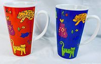Photo of two tall cat-themed mugs, one with a red background and one with a blue background, showing colorful cat illustrations and other small animals and plants