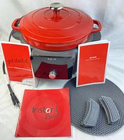 Red Instant Pot cast iron Dutch oven on electric heating base with silicone handle covers, heat diffuser pad, recipe book, and safety booklet.