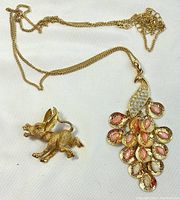 Photo showing the gold-tone donkey brooch with green eyes and peacock pendant on chain side by side on white background