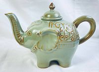 Side profile of ceramic elephant teapot showing trunk spout, ear, handle, and gold decorative accents.