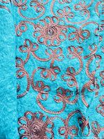Close-up photo showing the turquoise wool blend throw blanket with detailed dark brown embroidery in floral and swirl patterns.