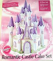 Box cover of Wilton Romantic Castle Cake Set showing a completed example of the 32 piece cake decorating set with white turrets and purple roofs and floral decorations.