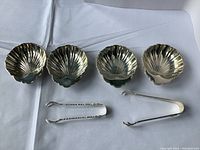 Four Birks sterling silver shell dishes and two sugar tongs arranged on a white cloth background.