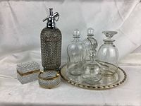 Photo showing collection of four glass decanters with stoppers on silver-tone tray, tall etched soda siphon, cut glass ashtray and cigarette box.