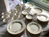 View of entire 67-piece Birks Grosvenor fine china dinnerware set arranged on table by window, showing dinner plates, salad plates, bread plates, dessert plates, teacups, saucers, teapot, sugar bowl, creamer, pedestal compote, and serving platters.