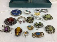 Top view of the full collection of 17 vintage jewelry pins and brooches laid on white cloth with measurements in background.