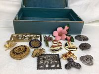 The lot of 14 vintage pins and brooches arranged in front of a blue box, showing various designs including floral, sailboats with enamel, and filigree metalwork.