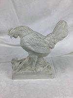 Side profile of white ceramic rooster figurine on rectangular base with chick.