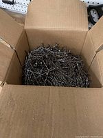 Open cardboard box filled with a large quantity of metal nails approximately 3.25 inches in length.