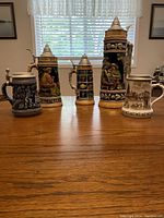 5 beer steins of varying heights displayed in a row on a table, showing general size comparison and designs