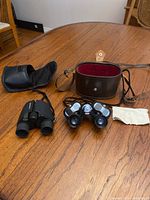 Two pairs of binoculars displayed on wood table with brown leather case, black pouch, and cleaning cloth.