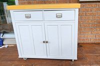 Front view showing two drawers with metal handles and two vertically paneled doors with round knobs, on caster wheels.