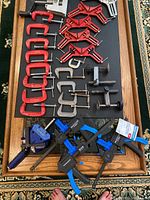 Photo showing an assortment of red and gray metal C-clamps of various sizes, small black C-clamps, black and blue quick-release bar clamps, and a blue small corner clamp arranged on a tray.