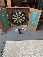 Wide view of dart board in wooden cabinet with open doors showing chalkboard scoreboards and dart accessories below