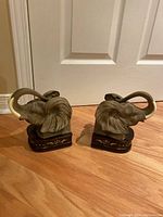 Front view of two elephant bust bookends showing detailed ears and trunks, wooden bases with gold paint, placed on wooden floor against white door.