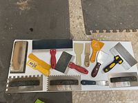 Full overhead view of assorted grout and tiling tools arranged on a white surface including floats, scrapers, brushes, and putty knives