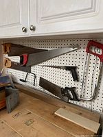 Full view of seven assorted hand saws hanging on pegboard including handsaws with wood and plastic handles and metal-frame hacksaws.