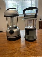 Front view of two LED lanterns placed side by side on wooden table in front of window with blinds. Lantern on left is Master Charge with a black dial and taller, lantern on right is Dorcy with handle and push button.