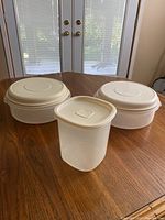 Overview of 3 plastic containers, 2 round and 1 rectangular, on wooden table near window.