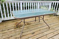 Outdoor rectangular glass top table with metal frame and umbrella hole, on wooden deck.