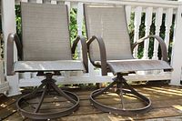 Two outdoor swivel chairs placed side-by-side on a wooden deck with white railing. Photo shows both chairs from a slightly angled front view highlighting the mesh fabric on the seats and backs, bronze circular swivel base, and armrests.
