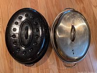 Top view of a black enamel roasting pan with raised round bumps on lid, next to a stainless steel roasting pan with vented lid and handles on wooden floor.