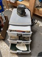 Three stacked plastic drawers with various sandpaper sheets and sanding accessories inside them