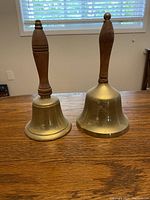 Front view of two brass school hand bells with wooden handles on a table, showing size and shape differences.
