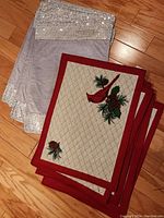 3 red cardinal embroidered quilted placemats stacked with 3 silver sequined border placemats underneath on hardwood floor