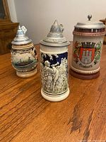 Front view of three covered German-style ceramic beer steins with metal lids placed on a wood table.