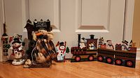 Wide view showing multiple Christmas holiday decorations including snowmen and a wooden train, placed against a white door and wooden floor.