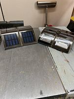 Four stainless steel solar lights shown together on a metal surface. Two lights have top-facing blue solar panels with clear covers, two have similar design with slightly different style.