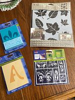 Photo of assorted stencils on table showing large leaf stencil, blue fleur-de-lis stencil, alphabet stencil 'A', and dark blue decorative stencil card all in original packaging.
