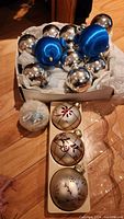Photo of assorted Christmas tree ornaments in a box on a wooden floor showing silver glass balls, blue plastic balls, cream and pink hand-painted glass balls, and a hand-painted candle.
